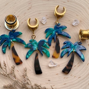 May include: Four pairs of gold palm tree earrings. Each earring features a blue and green palm tree with a brown trunk. The earrings are designed to be worn as ear gauges or plugs.