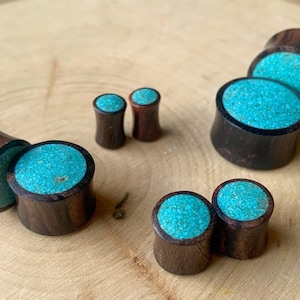 May include: Set of six wooden ear gauges with turquoise inlay. The gauges are different sizes and are arranged on a wooden surface.
