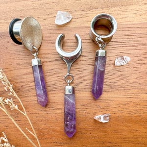 May include: Three silver ear gauges with amethyst stone pendants. The gauges are in the shape of a crescent moon, a circle, and a teardrop. The amethyst stones are a light purple color.