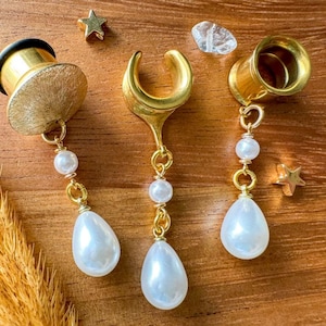 May include: Gold-coloured ear gauges with dangling white teardrop-shaped pearls and smaller white pearls. The gauges are displayed on a wooden surface, with gold star-shaped charms and a clear crystal. The jewellery has a celestial theme.