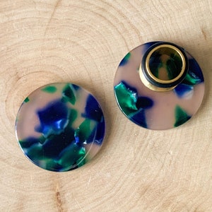 May include: Two round, brown, blue, and green patterned acrylic ear gauges. The larger gauge has a gold metal ring in the center.