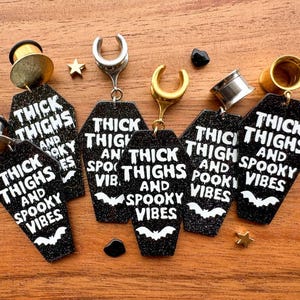 May include: Six black and white coffin-shaped earrings with the text "Thick Thighs and Spooky Vibes" on each. The earrings are made of acrylic and have a glittery finish.
