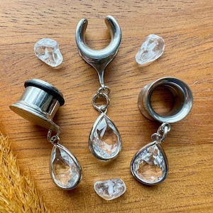 Puede incluir: Tres expansores de oreja plateados con colgantes de cristal transparente en forma de lágrima. Los expansores están hechos de acero quirúrgico y están disponibles en una variedad de tamaños.