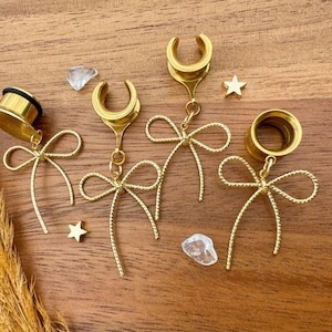 Puede incluir: Túneles dorados para las orejas con dijes en forma de lazo. Los túneles se exhiben sobre una superficie de madera, junto con pequeños elementos decorativos en forma de estrella y cristal. Diseñados para perforaciones de oreja estiradas.