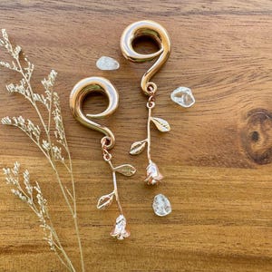 Puede incluir: Un par de dilatadores de oreja de oro rosa con colgantes de rosa y hoja. Los dilatadores están hechos de metal y tienen un acabado liso y pulido.