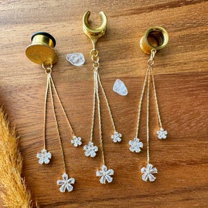 May include: Gold-coloured ear gauges with dangling chains decorated with sparkling, clear flower-shaped charms. The gauges are displayed on a wooden surface, showcasing their intricate design and elegant aesthetic. The jewellery is designed for stretched ear piercings.