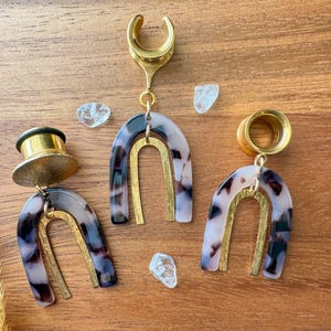 May include: Three gold and tortoise shell ear gauges with a gold arch design. The gauges are hanging from a wooden surface.