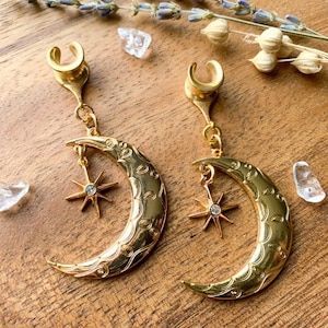 May include: A pair of gold crescent moon earrings with a star charm. The earrings are made of metal and have a detailed design.