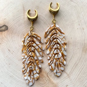 May include: A pair of gold ear gauges with white and gold feather-shaped dangles. The feathers have a marbled pattern.