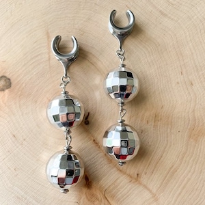 May include: A pair of silver earrings with a disco ball design. Each earring features a silver hoop with a silver chain attached to three silver disco ball-shaped beads.