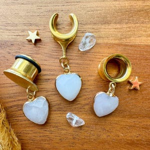 May include: Three gold ear gauges with white heart-shaped charms. The gauges are made of metal and have a shiny finish. The charms are made of stone and have a natural, cloudy appearance.