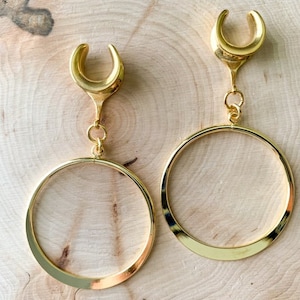 May include: A pair of gold-toned hoop earrings with a crescent moon-shaped dangle. The hoops are thick and have a smooth, polished finish.
