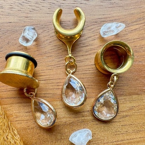 Puede incluir: Tres expansores de oreja dorados con dijes de cristal en forma de lágrima. Los expansores están hechos de metal y tienen un acabado brillante. Los dijes son transparentes y tienen un diseño facetado.