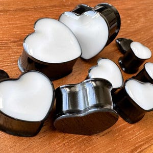 Pode incluir: Uma coleção de alargadores de orelha em forma de coração em preto e branco. Os alargadores têm um exterior preto brilhante e uma face branca em forma de coração. Os alargadores estão em vários tamanhos e são exibidos em uma superfície de madeira.