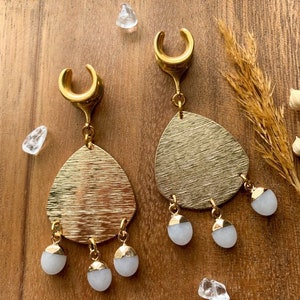 May include: A pair of gold ear cuffs with dangling teardrop-shaped charms. The charms are textured gold and have three white stone accents hanging from each.