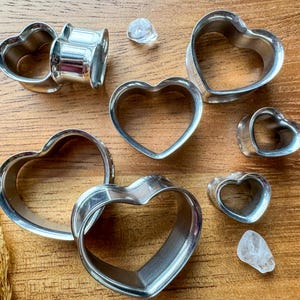 May include: An assortment of silver heart-shaped plugs and tunnels, of different sizes, displayed on a wooden surface. Two clear, irregularly shaped stones are also visible. These are body modification jewellery.