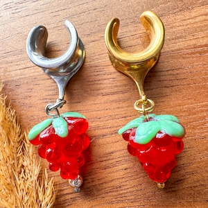 May include: A pair of ear gauges with a silver and gold finish. Each gauge features a dangling red strawberry charm with green leaves. The gauges are displayed on a wooden surface.