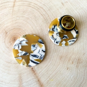 May include: Two round, gold, black, and white patterned acrylic ear gauges. One gauge is on a wooden surface and the other is on top of a gold metal ear gauge tunnel.