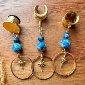 May include: Three gold-toned ear plugs featuring dangling charms. Each plug has a small blue bead, a larger marbled blue and turquoise stone bead, and a gold-toned starburst charm suspended within a thin gold-toned circular metal hoop frame.