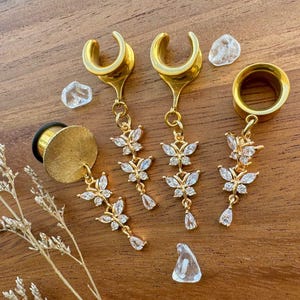 Puede incluir: Calibradores y túneles para orejas dorados con dijes de mariposa y cristal de lágrima. Las joyas se exhiben sobre una superficie de madera con pequeñas piedras decorativas transparentes. Diseñado para piercings de oreja estirados.
