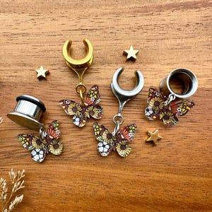 May include: Three pairs of butterfly dangle earrings. Each pair features a silver or gold hoop with a dangling butterfly charm. The butterflies are colorful and have a floral pattern.