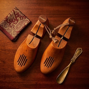 May include: A pair of light brown wooden shoe trees with the brand name Florsheim engraved on the heel knobs, accompanied by a dark red pocket square with a gold paisley pattern and a small, tarnished brass shoehorn with an ornate handle.