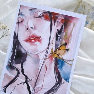 Puede incluir: Impresión artística en acuarela que presenta un retrato de una persona con los ojos cerrados, labios rojos y pelo largo y negro. Una mariposa con alas marrones y amarillas está cerca del cuello. La obra de arte tiene una calidad suave y etérea con acentos rojos y dorados.