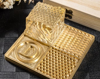 Könnte beinhalten: Ein goldfarbenes Tablett aus Metall mit vier verschiedenen strukturierten Abschnitten. Das Tablett hat einen erhabenen Buchstaben "D" im mittleren Abschnitt. Das Tablett liegt auf einer schwarzen strukturierten Oberfläche.