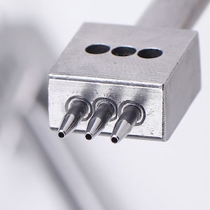 May include: A silver metal tool with three round holes and three pointed prongs. The tool is used for punching holes in leather or other materials.