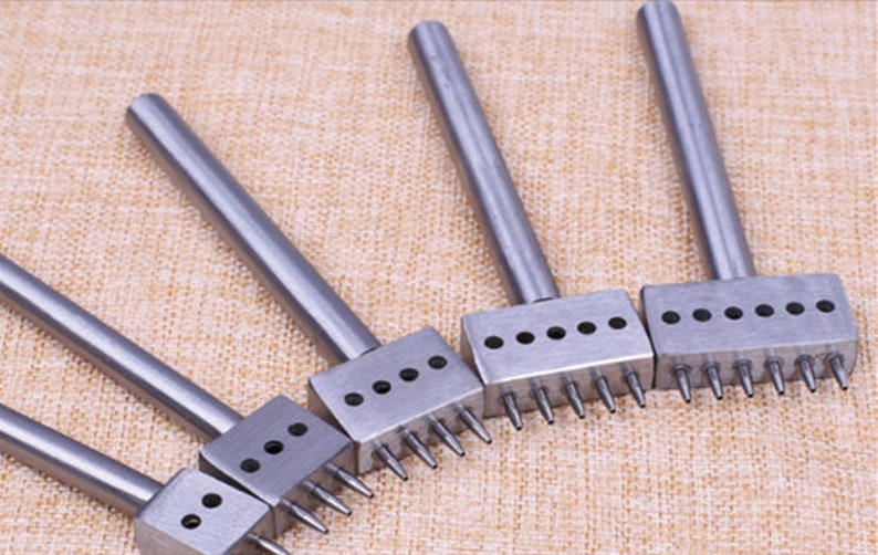 May include: Set of five metal leather hole punches with different numbers of holes. The punches have long handles and are arranged in a row on a beige fabric surface.