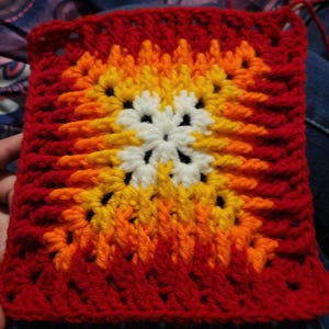 May include: A square crochet motif featuring a central white flower pattern surrounded by concentric layers of yarn in shades of yellow, orange, and deep red. The piece is constructed with a classic granny square stitch pattern, creating a textured, geometric design with visible yarn loops and open spaces throughout the square.