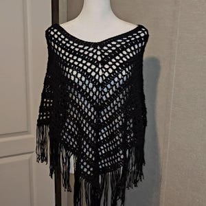 May include: A black crochet poncho featuring an open, diamond-shaped stitch pattern throughout the garment. The piece has a V-shaped hemline finished with long, dangling fringe trim along the bottom edges, creating a textured and layered appearance.