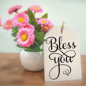May include: A white tag with black cursive lettering that says "Bless you" with a small heart symbol below the text. The tag is hanging from a bouquet of pink flowers in a white ceramic vase.