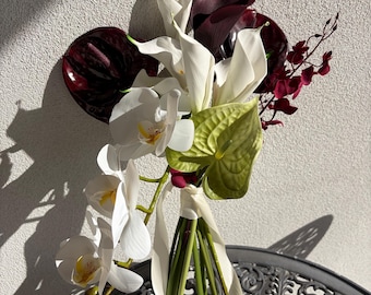May include: A floral bouquet featuring white calla lilies, white orchids with yellow centers, deep burgundy anthuriums, a light green anthurium, and small dark red accent flowers. The stems are bound together with a cream-colored ribbon.