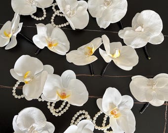 May include: A collection of artificial white orchid flowers, each featuring a yellow and pink centre. Several of the orchid stems are accented with loops of small white faux pearls, while others have simple black stems, arranged on a dark surface.