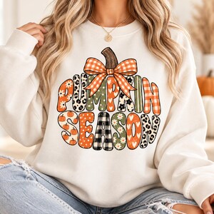 May include: A white crewneck sweatshirt featuring a large graphic of a pumpkin shape filled with various patterns including orange plaid, leopard print, white daisies, and black buffalo check, with the text PUMPKIN SEASON and an orange plaid bow.