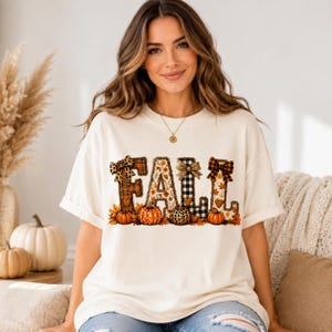 Op de afbeelding: Een crèmekleurig T-shirt met korte mouwen en het woord FALL in grote blokletters. Elke letter heeft een uniek patroon, waaronder ruitjes, luipaardprint en herfstbladeren. Kleine oranje pompoenen en strikjes zijn in het ontwerp verwerkt.