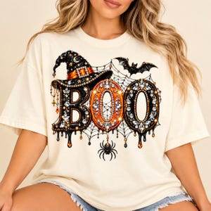 Op de afbeelding: Een crèmekleurig T-shirt met korte mouwen en een grafische print van het woord BOO in zwarte en oranje letters met juwelen. Het ontwerp bevat een heksenhoed, een spinnenweb, een hangende spin en een vleermuis met sprankelende accenten.
