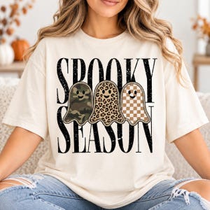 Op de afbeelding: Een crèmekleurig t-shirt met de zwarte tekst SPOOKY SEASON in een verweerd lettertype. Onder de tekst staan drie spookillustraties, elk met een ander patroon: één in groen camouflage, één in luipaardprint en één in een beige dambordpatroon.