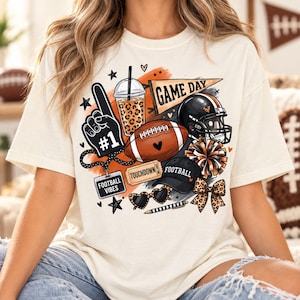Puede incluir: Una camiseta color crema con un estampado gráfico de artículos de fútbol americano, incluyendo un dedo de espuma número uno, una bebida con estampado de leopardo, un banderín que dice GAME DAY, un balón, un casco, un boleto y una gorra.