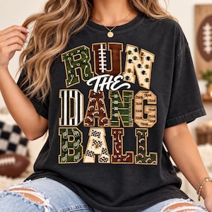 Op de afbeelding: Een antracietgrijs t-shirt met korte mouwen en de tekst RUN THE DANG BALL in grote blokletters. Elke letter is gevuld met diverse patronen in het thema American football, waaronder spelschema's, footballs, helmen en veldlijnen.