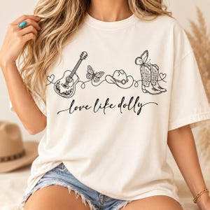Könnte beinhalten: Ein cremefarbenes T-Shirt mit einer schwarzen Strichgrafik, die eine akustische Gitarre, zwei Schmetterlinge, einen Cowboyhut und ein Paar Cowboystiefel zeigt. Der Text "love like dolly" ist in schwarzer Schreibschrift darunter gedruckt.