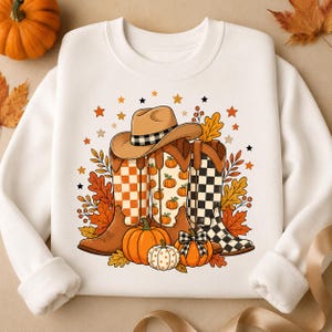 Può includere: Una felpa bianca a girocollo con una stampa grafica raffigurante tre stivali da cowboy e un cappello da cowboy. Gli stivali presentano motivi a scacchi arancioni e neri e disegni di zucche. Foglie autunnali arancioni, zucche e piccole stelle circondano gli stivali.