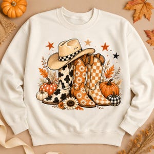 Könnte beinhalten: Ein cremefarbenes Sweatshirt mit Rundhalsausschnitt und einem Grafikdruck eines Cowboyhuts, der auf drei Cowboystiefeln ruht. Die Stiefel zeigen Kuhflecken-, Gänseblümchen- und Karomuster, umgeben von orangefarbenen Kürbissen, Herbstblättern und Sternen.