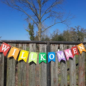 Könnte beinhalten: Ein farbenfrohes Banner mit dem Wort "WILLKOMMEN" in weißen Buchstaben auf einem Holzzaun. Das Banner besteht aus einzelnen Flaggen in verschiedenen Farben des Regenbogens.
