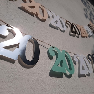Könnte beinhalten: Ein dekoratives Banner mit den Zahlen "2020" in verschiedenen Farben, darunter Silber, Grün und Beige. Die Zahlen sind auf einer dünnen Schnur aufgereiht, perfekt für eine Abschlussfeier oder Neujahrsfeier.