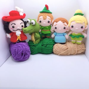 Puede incluir: Juguetes de amigurumi tejidos a crochet de Peter Pan, el Capitán Garfio, Campanita y un cocodrilo. Los juguetes están dispuestos en fila con ovillos de lana delante de ellos.