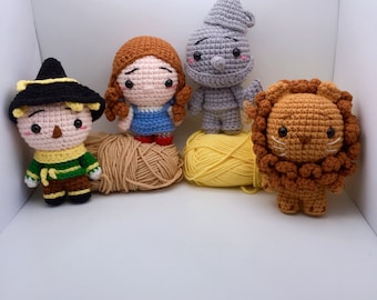 May include: Crocheted stuffed toys of Dorothy, the Scarecrow, the Tin Man, and the Cowardly Lion from the Wizard of Oz. The toys are sitting on a white surface with yarn balls.
