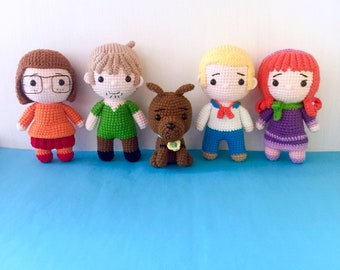 May include: Five crocheted characters from the cartoon Scooby-Doo. The characters are Velma, Shaggy, Fred, Daphne, and Scooby-Doo. The characters are all standing on a blue surface.