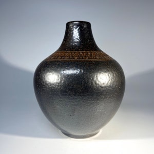 Puede incluir: Un jarrón de cerámica gris oscuro con un diseño geométrico marrón alrededor del centro. El jarrón tiene un cuerpo redondeado y un cuello estrecho.